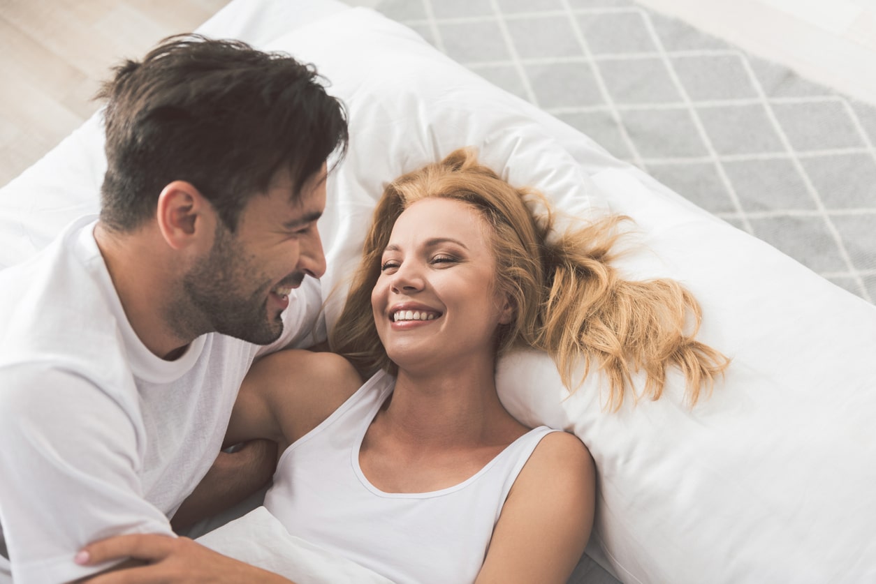 A younger white couple reclines on a bed, smiling and laughing in white shirts