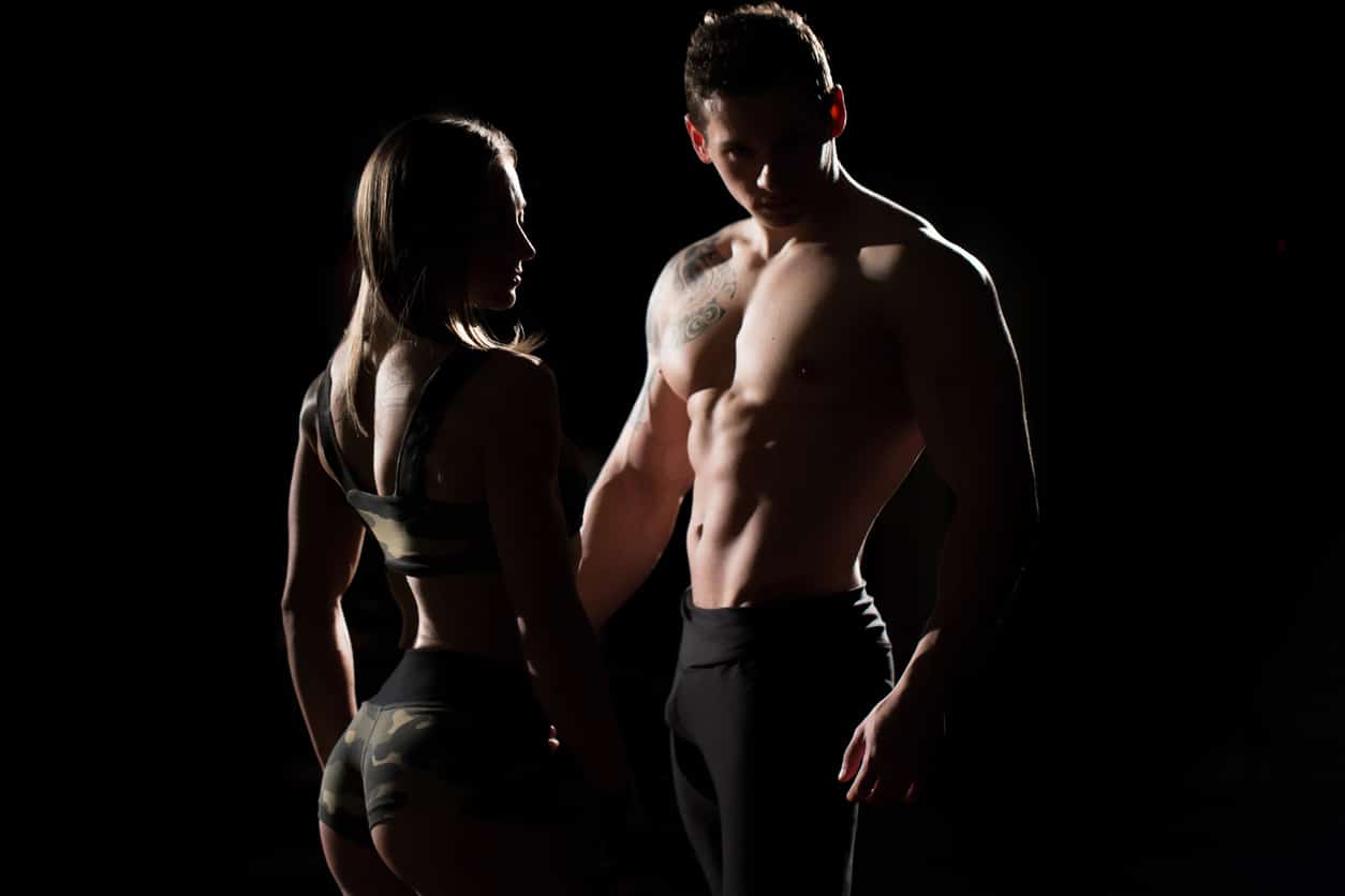 A muscular man and woman pose in shadow while light accentuates their sculpted bodies