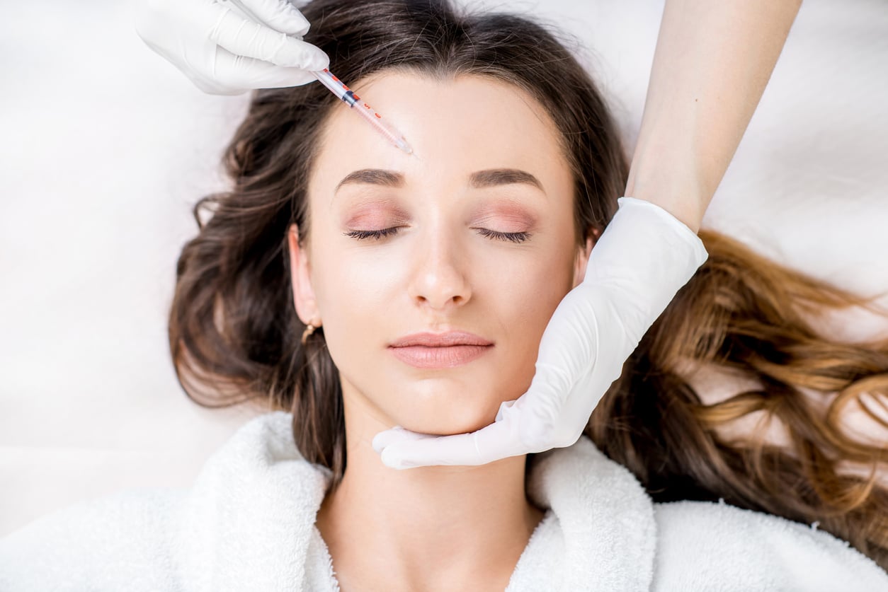 A brunette woman with flowing hair relaxes with her eyes closed while glove hands position a Botox injection